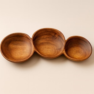 May include: A wooden serving bowl with three connected circular compartments. The bowl is made of a light brown wood with visible grain patterns. The design is ideal for serving snacks or appetizers.