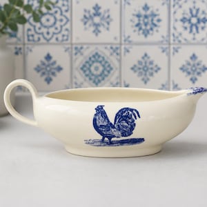 May include: A cream-colored ceramic gravy boat featuring a blue rooster illustration on the side. The vessel has a single curved handle on one side and a pouring spout on the other, with a blue decorative accent on the rim of the spout.