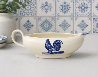 May include: A cream-coloured ceramic gravy boat featuring a blue rooster illustration on the side. The vessel has a single curved handle on one side and a pouring spout on the other, with a blue decorative accent on the rim of the spout.