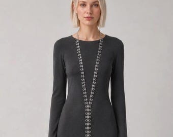May include: A long silver-toned necklace featuring a series of interconnected circular links that form a vertical chain, worn over a dark gray long-sleeved dress with a round neckline. The necklace extends down the center of the torso.