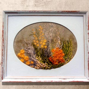 A close-up shot of a hand-embroidered oval floral meadow study with yellow and orange wool yarn sunburst blooms on a natural linen weave ground, presented inside a distressed light blue wooden frame hanging directly on a textured, light gray concrete-look wall.