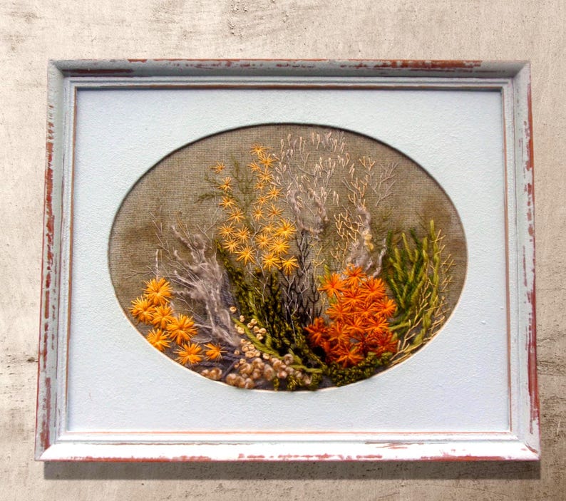 A close-up shot of a hand-embroidered oval floral meadow study with yellow and orange wool yarn sunburst blooms on a natural linen weave ground, presented inside a distressed light blue wooden frame hanging directly on a textured, light gray concrete-look wall.