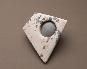 May include: A diamond-shaped, off-white stone object with gray and brown speckles. A circular, gray metallic element is set into the center. The object is likely a decorative element or a piece of art.