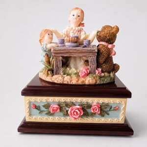 May include: A decorative music box featuring a sculpted scene of two figures and a brown teddy bear sitting at a wooden table. The base is dark wood with a light blue panel decorated with pink roses and green leaves in a relief pattern.