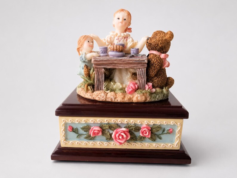 May include: A decorative music box featuring a sculpted scene of two figures and a brown teddy bear sitting at a wooden table. The base is dark wood with a light blue panel decorated with pink roses and green leaves in a relief pattern.