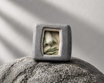 May include: A gray, cube-shaped sculpture with a carved face in a rectangular opening. The face has green and off-white tones, with a prominent nose and lips. The sculpture rests on a gray, textured rock.