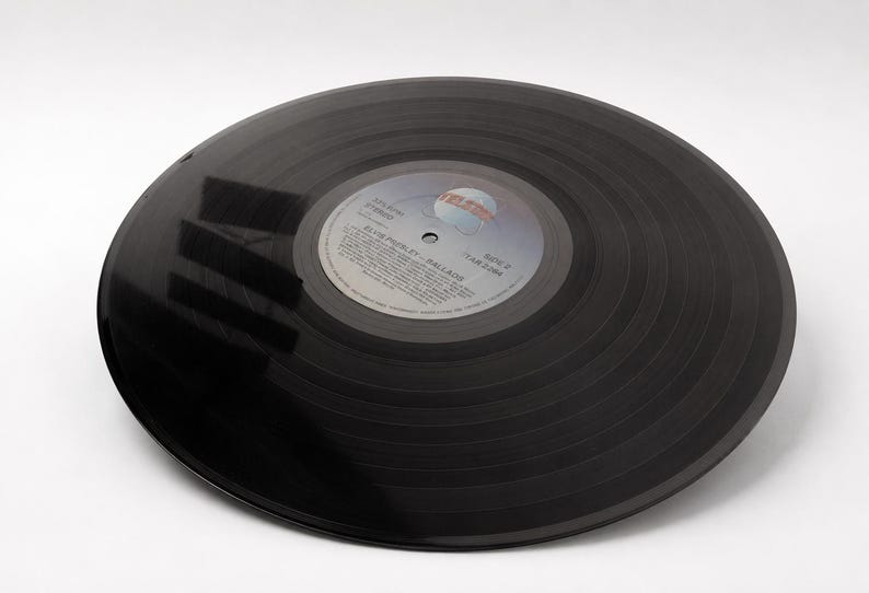 A black 12 inch vinyl record lies flat on a white surface with clear dark reflections across its grooved surface.
