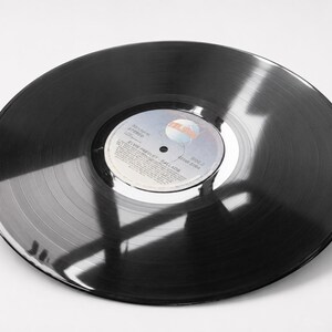 A black 12 inch vinyl record lies flat on a white surface with light reflecting across its grooved surface.