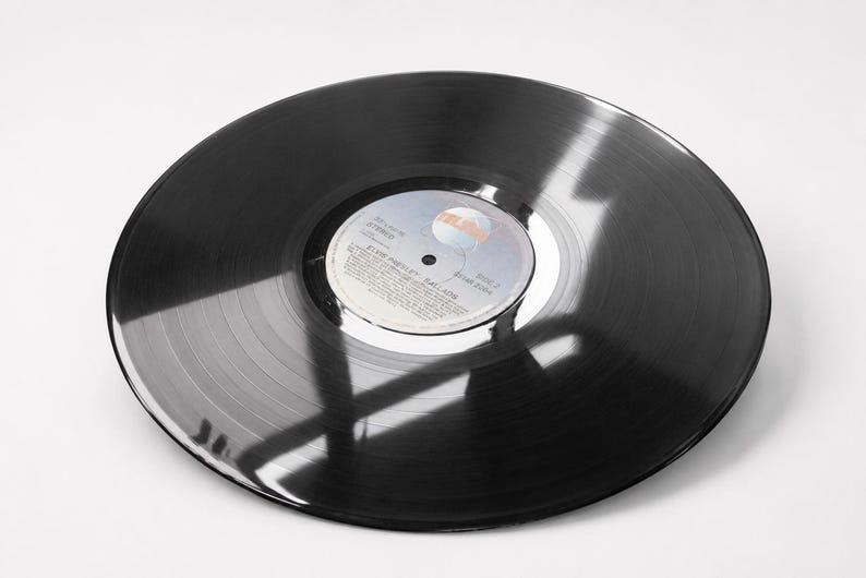 A black 12 inch vinyl record lies flat on a white surface with light reflecting across its grooved surface.