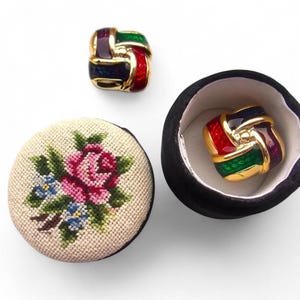 May include: A set of vintage earrings and a small round container. The earrings are gold-toned with red, green, purple, and blue enamel. A small, round, cross-stitched container with a floral design is also present.