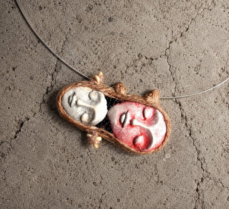 Two clay faces hand-shaped into an oval pendant bound by brown twine on a silver wire choker.