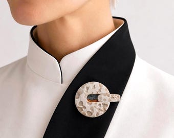 May include: A circular brooch with a textured, off-white surface and a small, rectangular accent. The brooch is attached to a black and white garment. The design is modern and minimalist, suitable for adding a touch of elegance to any outfit.