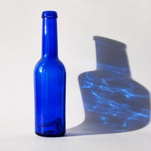 Vintage cobalt blue glass bottle standing upright beside its dramatic blue glass shadow with rippling light reflections, photographed against a clean light grey-white studio background.
