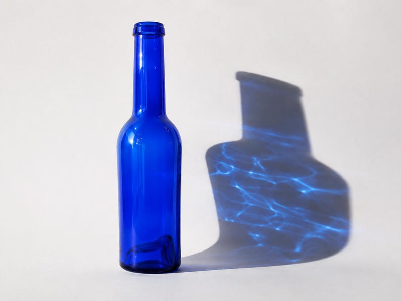 Vintage cobalt blue glass bottle standing upright beside its dramatic blue glass shadow with rippling light reflections, photographed against a clean light grey-white studio background.