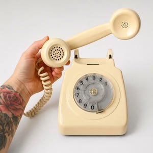 A hand with a rose tattoo holding a cream-colored rotary dial telephone receiver high above its matching base station against a plain white background.
