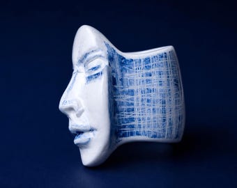 May include: A white ceramic ring featuring a sculpted human face in profile on the front. The surface is decorated with blue painted accents and a cross-hatched pattern that extends across the side of the band, creating a textured appearance.