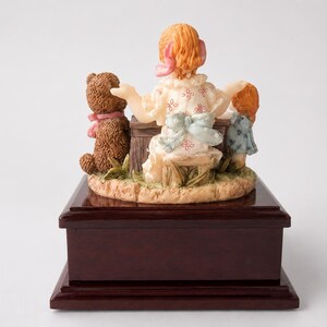 May include: A decorative figurine featuring a central figure with reddish hair and a white dress with pink patterns, seated on a wooden stool. Beside the figure are a brown teddy bear with a pink ribbon and a smaller figure in a blue dress.