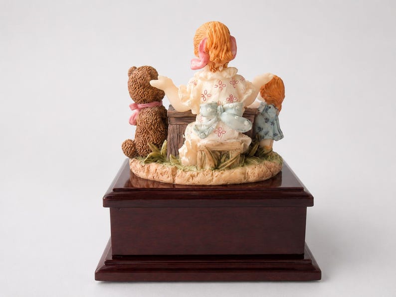 May include: A decorative figurine featuring a central figure with reddish hair and a white dress with pink patterns, seated on a wooden stool. Beside the figure are a brown teddy bear with a pink ribbon and a smaller figure in a blue dress.