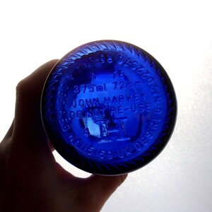Close-up of the cobalt blue glass bottle base held in a hand, showing raised embossed markings including “25,” “375 ml 72 mm,” “JOHN HARVEY & SONS,” “VINO NON DISPERSE,” “19,” and “3,” with a raised central emblem and circular embossed lettering around the rim in a light grey studio.