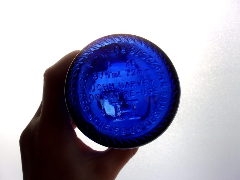 Close-up of the cobalt blue glass bottle base held in a hand, showing raised embossed markings including “25,” “375 ml 72 mm,” “JOHN HARVEY & SONS,” “VINO NON DISPERSE,” “19,” and “3,” with a raised central emblem and circular embossed lettering around the rim in a light grey studio.