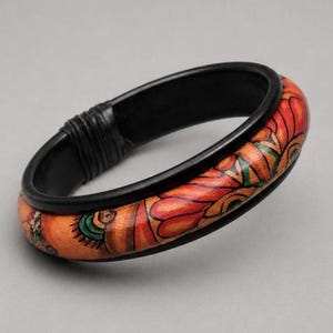 May include: A bangle bracelet featuring a smooth black inner band and a contrasting outer band decorated with a vibrant orange, red, and green floral pattern. The bracelet includes a section wrapped in black cord, providing a textured detail.