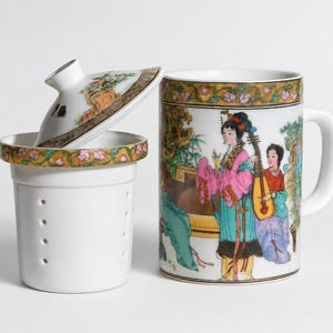White porcelain three piece tea set including a straight sided mug with a handle decorated with a colorful illustration of asian women playing musical instruments in a garden a matching white ceramic infuser with drainage holes and a round white porcelain lid with floral borders on a plain white background.