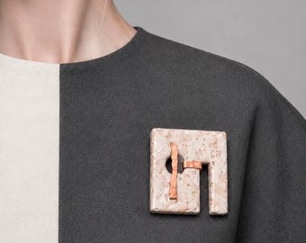 May include: A geometric brooch with a square, speckled, off-white base and a copper-colored accent. The brooch is attached to a dark gray garment, with a white panel visible on the left side. The jewelry piece has a modern, minimalist design.