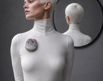 May include: A white long-sleeve turtleneck shirt featuring a textured, round brooch with a mottled grey and pink surface. The brooch has a row of small circular indentations along the bottom edge, pinned to the chest of the garment.
