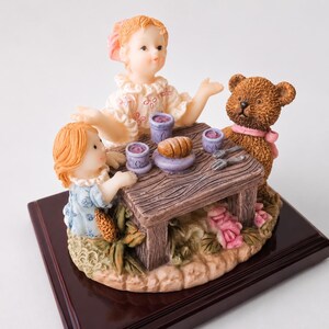 May include: A decorative figurine featuring two children and a brown teddy bear sitting at a small wooden table. The table holds three purple cups and a loaf of bread. The figures are set on a base with sculpted pink flowers and green leaves.