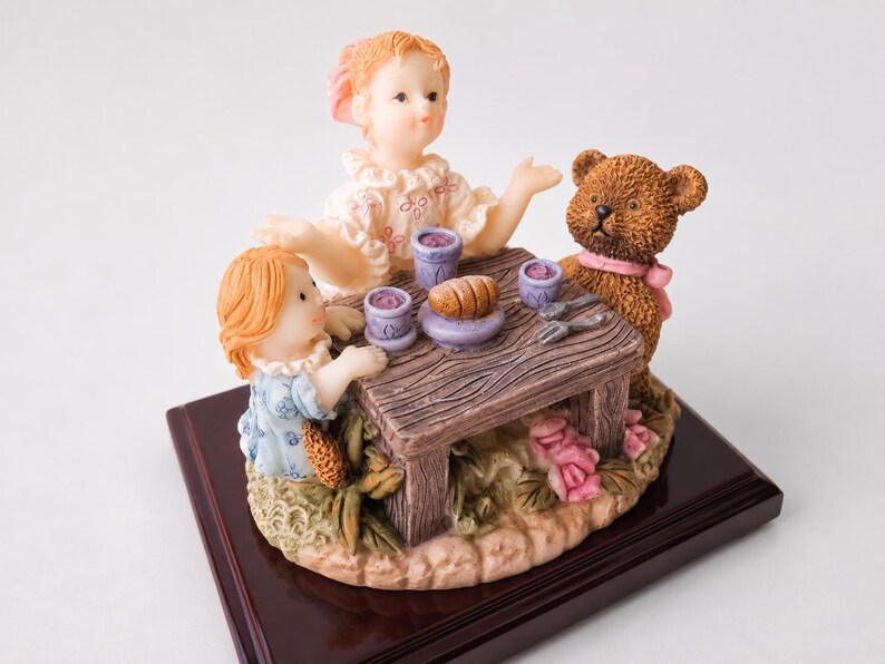 May include: A decorative figurine featuring two children and a brown teddy bear sitting at a small wooden table. The table holds three purple cups and a loaf of bread. The figures are set on a base with sculpted pink flowers and green leaves.