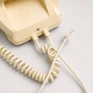 A close-up view of the back panel of a cream-colored telephone showing the coiled handset cord and a straight line cord with a clear plastic modular plug against a white background.