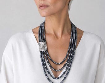 May include: A multi-strand necklace featuring four rows of dark gray spherical beads. The strands are connected on one side by a rectangular silver-toned clasp embellished with clear, sparkling stones arranged in a geometric grid pattern.