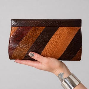 Pode incluir: Uma clutch marrom e laranja com um padrão de listras diagonais. A clutch é feita de couro texturizado e tem formato retangular com a parte superior curva. A paleta de cores inclui tons de marrom e laranja.
