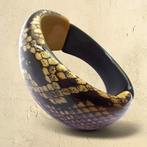 May include: A patterned bangle bracelet with a snake-like design. The bracelet has a black interior and a tan accent piece. The exterior has a pattern of tan and brown scales. A bold fashion accessory.