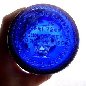 Close-up of the cobalt blue glass bottle base held in a hand, showing raised embossed markings including “25,” “375 ml 72 mm,” “JOHN HARVEY & SONS,” “VINO NON DISPERSE,” “19,” and “3,” with a raised central emblem and circular embossed lettering around the rim.