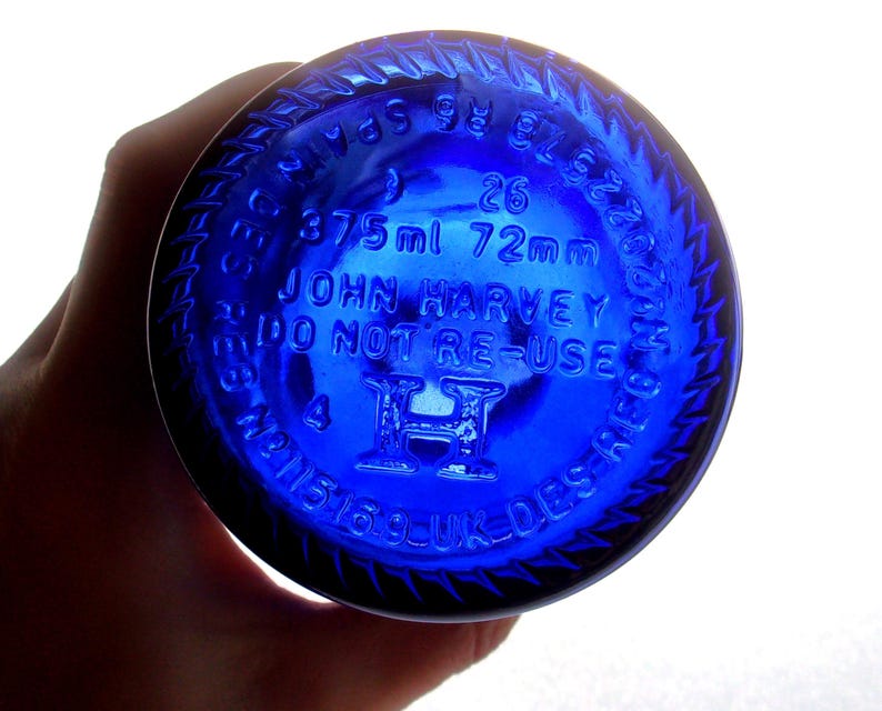 Close-up of the cobalt blue glass bottle base held in a hand, showing raised embossed markings including “25,” “375 ml 72 mm,” “JOHN HARVEY & SONS,” “VINO NON DISPERSE,” “19,” and “3,” with a raised central emblem and circular embossed lettering around the rim.