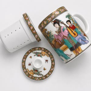 White porcelain three piece tea set including a straight sided mug with a handle decorated with a colorful illustration of asian women playing musical instruments in a garden a matching white ceramic infuser with drainage holes and a round white porcelain lid with floral borders on a plain white background.