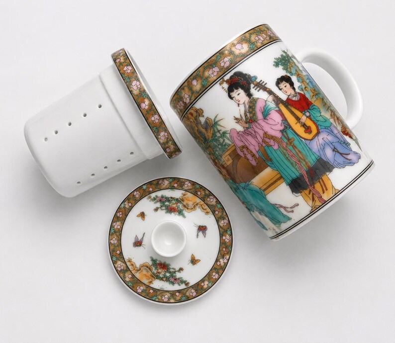 White porcelain three piece tea set including a straight sided mug with a handle decorated with a colorful illustration of asian women playing musical instruments in a garden a matching white ceramic infuser with drainage holes and a round white porcelain lid with floral borders on a plain white background.