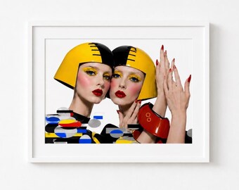 May include: A framed art print featuring two individuals wearing matching yellow and black helmets with bold makeup, including yellow eyeshadow and red lipstick. The composition includes abstract geometric shapes in blue, yellow, and black.