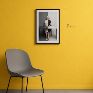 A framed A4 art print displayed on a vibrant yellow interior wall. The artwork features a minimalist portrait of a seated woman in a monochrome black-and-white composition, presented within a sleek black frame. A stylish grey upholstered accent chair sits nearby, creating a modern Scandinavian-inspired interior that highlights the scale of the A4 print.