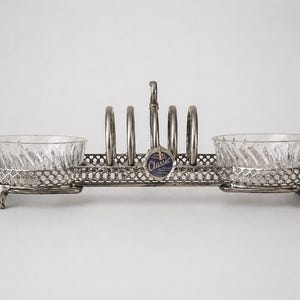 May include: An antique silver-toned toast rack with two clear glass bowls. The rack has a decorative openwork design and four vertical loops for holding toast. A small tag with the word "Classic" hangs from the top.