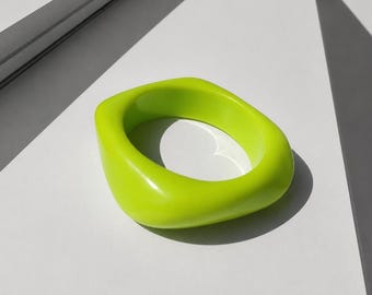 May include: A bright lime green bangle bracelet with a smooth, glossy finish and a rounded square shape. The accessory features a thick, solid construction with a wide band and a circular opening in the center, showcasing a vibrant, solid color.