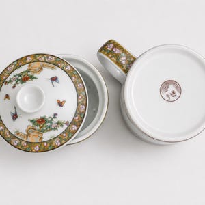 White porcelain three piece tea set including a straight sided mug with a handle decorated with a colorful illustration of asian women playing musical instruments in a garden a matching white ceramic infuser with drainage holes and a round white porcelain lid with floral borders on a plain white background.