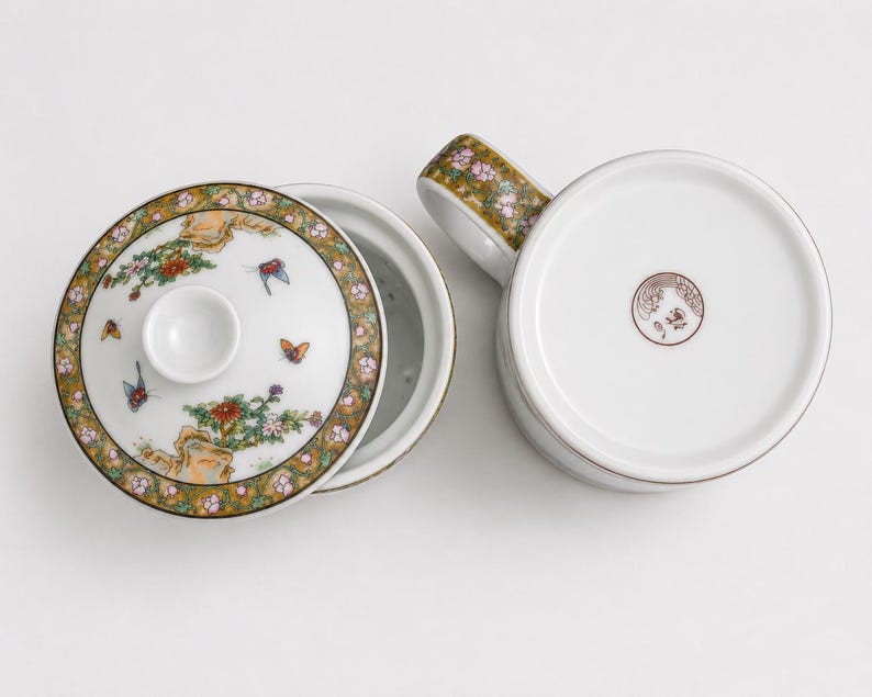 White porcelain three piece tea set including a straight sided mug with a handle decorated with a colorful illustration of asian women playing musical instruments in a garden a matching white ceramic infuser with drainage holes and a round white porcelain lid with floral borders on a plain white background.