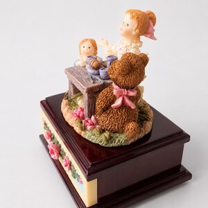 May include: A music box featuring a dark brown wooden base with pink rose accents. On top, a figurine depicts a child with light hair and a pink bow, a smaller doll, and a brown teddy bear with a pink ribbon sitting at a small wooden table.