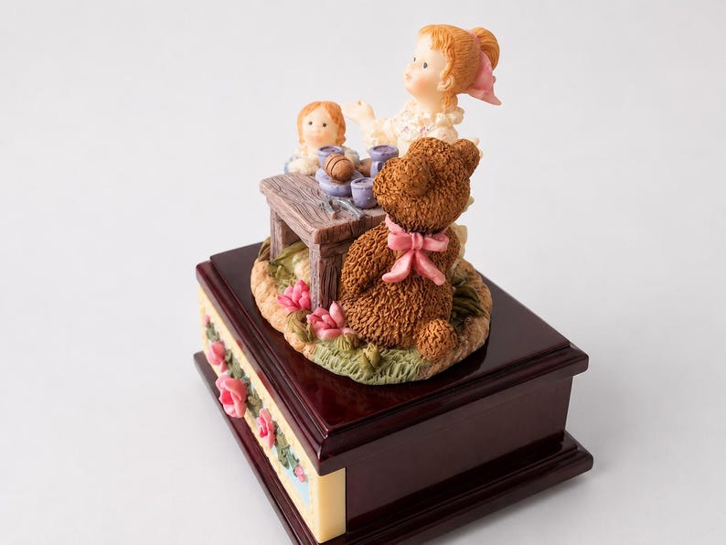 May include: A music box featuring a dark brown wooden base with pink rose accents. On top, a figurine depicts a child with light hair and a pink bow, a smaller doll, and a brown teddy bear with a pink ribbon sitting at a small wooden table.
