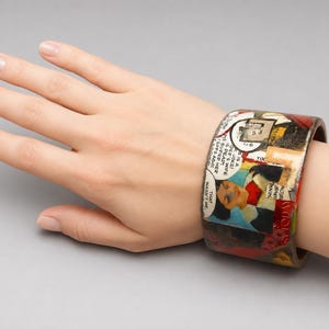 Handmade Comic Book Pop Art bangle worn on a hand, showing its wide upcycled wood cuff design with vintage comic panels, speech bubbles, illustrated figures and bold red, blue, black and cream colours.