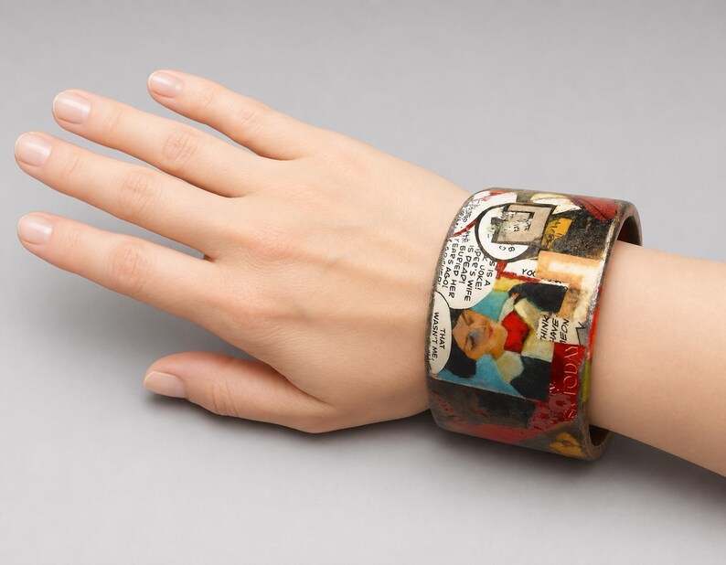 Handmade Comic Book Pop Art bangle worn on a hand, showing its wide upcycled wood cuff design with vintage comic panels, speech bubbles, illustrated figures and bold red, blue, black and cream colours.