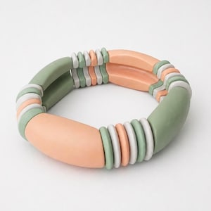 May include: A multi-coloured bracelet with peach, sage green, and white beads. The bracelet has a chunky design with rounded, oblong beads and smaller, circular beads. The colour palette is soft and neutral, suitable for everyday wear.