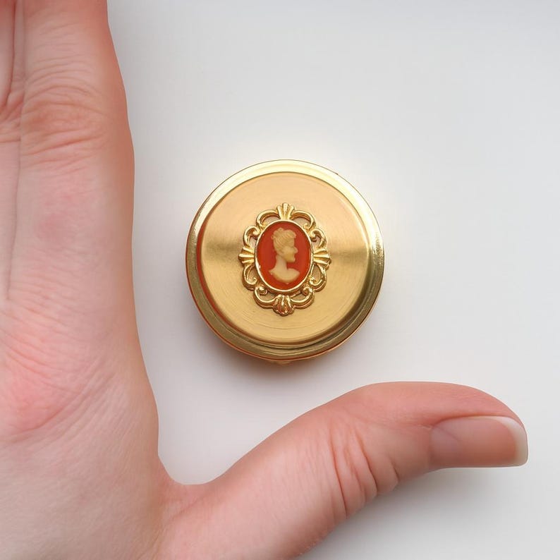 A small vintage pillbox with a red-and-ivory cameo trinket framed in gold filigree, this gold keepsake doubles as a mini treasure and handmade box. Styled as a regency charm, it’s perfect for gifting as a victorian gift or historical gift. The aged finish enhances its appeal as an antique case, ideal for retro cottage collectors seeking micro luxury and collectible art in a timeless rose gold box.